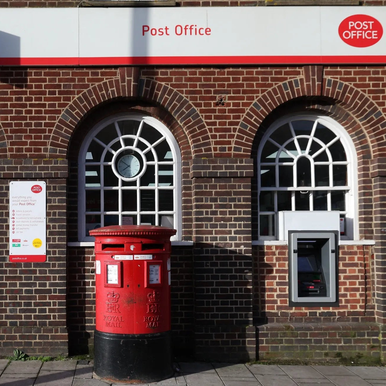 Red post box in front of a brick building with 'Post Office' sign.